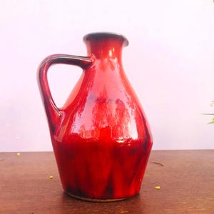May include: A ceramic pitcher with a glossy, vibrant red glaze that transitions into darker, deep burgundy tones near the base. The vessel features a rounded body, a narrow neck with a flared rim, and a single curved handle on the left side.