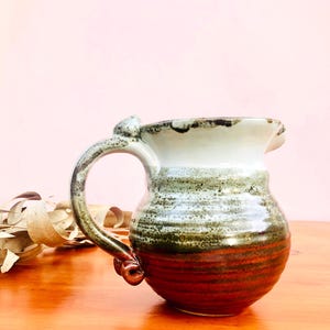 May include: A ceramic pitcher featuring a rounded body with a horizontal ribbed texture. The glaze transitions from a creamy white at the rim to a speckled sage green in the middle, ending in a deep reddish-brown base with a curved handle.