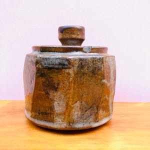 May include: A cylindrical ceramic jar with a matching lid, featuring a mottled glaze in shades of brown, tan, and dark grey. The vessel has a faceted, multi-sided exterior surface with a textured, earthy appearance and a small, knob-like handle.