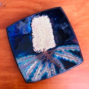 May include: A square ceramic tile featuring a glossy deep blue glaze with a central raised relief of a white banksia flower. The bottom section displays stylised green and blue foliage patterns with intricate textures and dark outlines.
