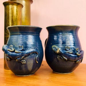 May include: Two dark blue ceramic mugs with a glossy finish, each featuring a raised, sculpted lizard relief on the exterior surface. The mugs have a rounded body shape with a wide rim and a smooth, deep blue glaze that shows subtle tonal variations.