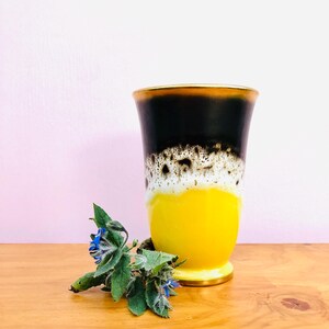 May include: A ceramic vase featuring a gradient colour scheme that transitions from a solid black top through a speckled white middle section to a solid yellow base. The rim and the base of the vase are finished with a thin gold-coloured trim.