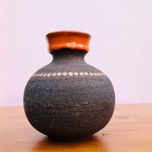 May include: A round ceramic vase with a textured dark brown body and a smooth, glossy orange rim. A horizontal band of small, light-coloured circular dots encircles the upper portion of the vase, contrasting with the rough, dark surface texture.