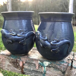 May include: Two dark blue ceramic mugs with a glossy finish, each featuring a raised, sculpted lizard relief wrapped around the exterior. The vessels have a rounded, bulbous shape with a wide rim and a smooth, uniform deep blue glaze throughout.