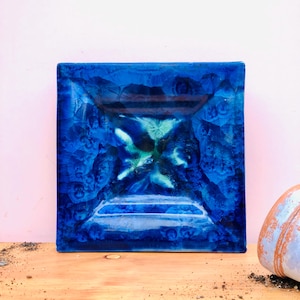 May include: A square glass dish featuring a deep cobalt blue glaze with crystalline patterns. The centre displays a starburst effect in shades of teal and white, while the edges show a darker, saturated blue hue with a smooth, glossy finish.