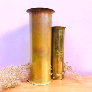May include: Two cylindrical brass shell casings of varying heights, both featuring a smooth, metallic finish with natural patina and flared rims. The taller casing stands to the left, while the shorter, slightly narrower casing is positioned to the right.