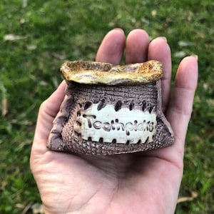 May include: A small, brown, textured ceramic container shaped like a rustic sack with a wide, open top. A rectangular, off-white patch is centred on the front, featuring the embossed, dark-coloured text "Toothpicks" in a clear, sans-serif font.