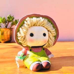 May include: A ceramic figurine of a child wearing a pink bonnet with a green interior and a green overall outfit. The figure has curly blonde yarn hair, black bead eyes, and rosy cheeks. One hand holds a small green leaf-shaped accessory.