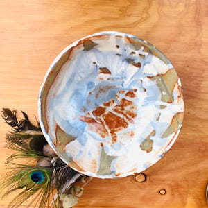 May include: A round ceramic bowl featuring an abstract glaze pattern in shades of white, light blue, and earthy brown. The interior surface displays a marbled effect with visible texture and irregular colour transitions across the curved basin.