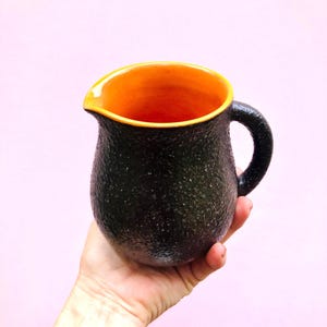 May include: A ceramic jug with a textured, dark charcoal-coloured exterior and a smooth, vibrant orange interior. The vessel features a rounded body, a curved handle on the side, and a small pouring spout at the top rim for liquid dispensing.