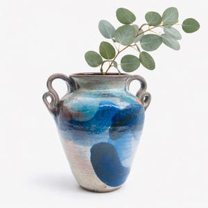 May include: A ceramic vase with a blue and brown abstract design, featuring two handles and a wide opening. The vase holds a sprig of eucalyptus with round, grey-green leaves. The vase is a decorative home decor item.