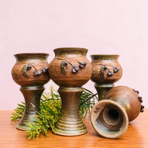 May include: Four brown ceramic goblets with a textured finish and a flared base. Each goblet features a relief design of small dark leaves and seed pods. One goblet is lying on its side, revealing the hollow interior and a small mark on the base.