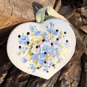 May include: A heart-shaped white ceramic ornament featuring a floral pattern of blue and yellow flowers with green leaves. The ornament has several small circular holes scattered across its surface and includes a light green ribbon for hanging.