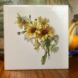 May include: A square white ceramic tile featuring a central illustration of a cluster of yellow and golden-brown daisies with green stems and leaves. The floral design is rendered in a painterly style with soft shading and visible brush strokes.