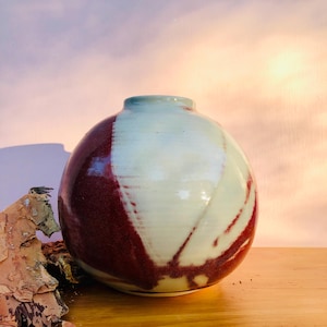 May include: A spherical ceramic vase featuring a glossy finish with a two-tone colour scheme. The surface displays a creamy off-white base colour contrasted by deep, rich maroon sections that wrap around the sides in a sweeping, organic pattern.