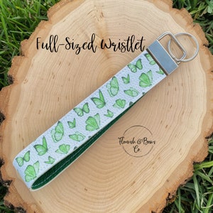 May include: Full-sized wristlet with a white background and green butterfly pattern. The wristlet has a silver key ring and a dark green interior. The text "Flourish & Bows Co." is visible.