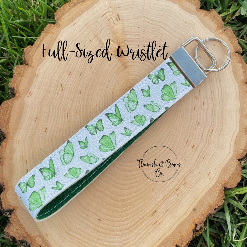 May include: Full-sized wristlet with a white background and green butterfly pattern. The wristlet has a silver key ring and a dark green interior. The text "Flourish & Bows Co." is visible.