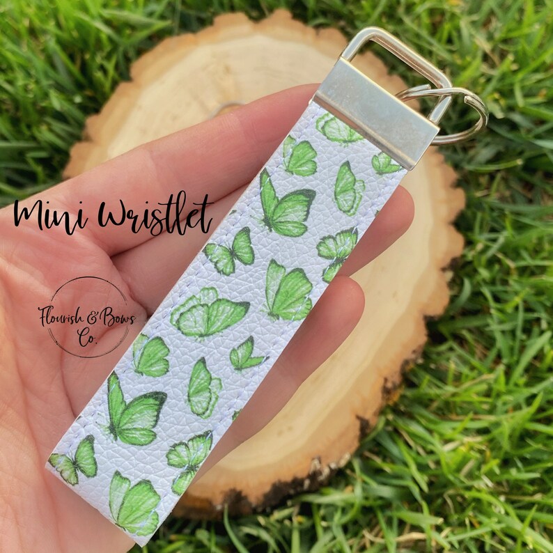 May include: A white mini wristlet with a pattern of green butterflies. The wristlet has a silver-colored metal clasp and a key ring. The text "Mini Wristlet" and "Flourish & Bows Co." are printed on the wristlet.