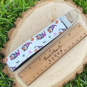 May include: White wristlet key fob with a Halloween gnome pattern. The key fob features a silver-tone metal ring and a black interior. A wooden ruler is next to the key fob, measuring in inches.