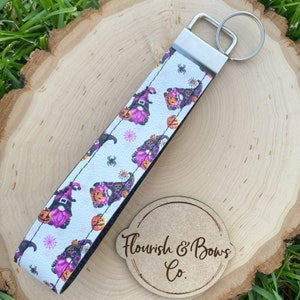 May include: A white wristlet keychain with a Halloween gnome pattern. The pattern includes purple gnomes, orange pumpkins, spiders, and spiderwebs. The keychain has a silver metal clasp and key ring. The brand name "Flourish & Bows Co." is visible.