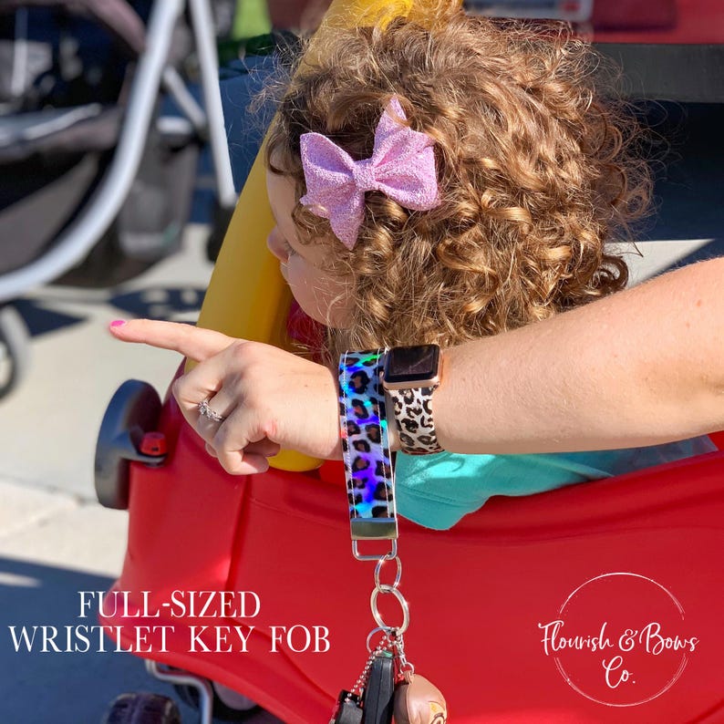 May include: A full-sized wristlet key fob with a colorful, iridescent leopard print design. The key fob is attached to a set of keys. The image also shows a smartwatch with a leopard print band and a pink bow.