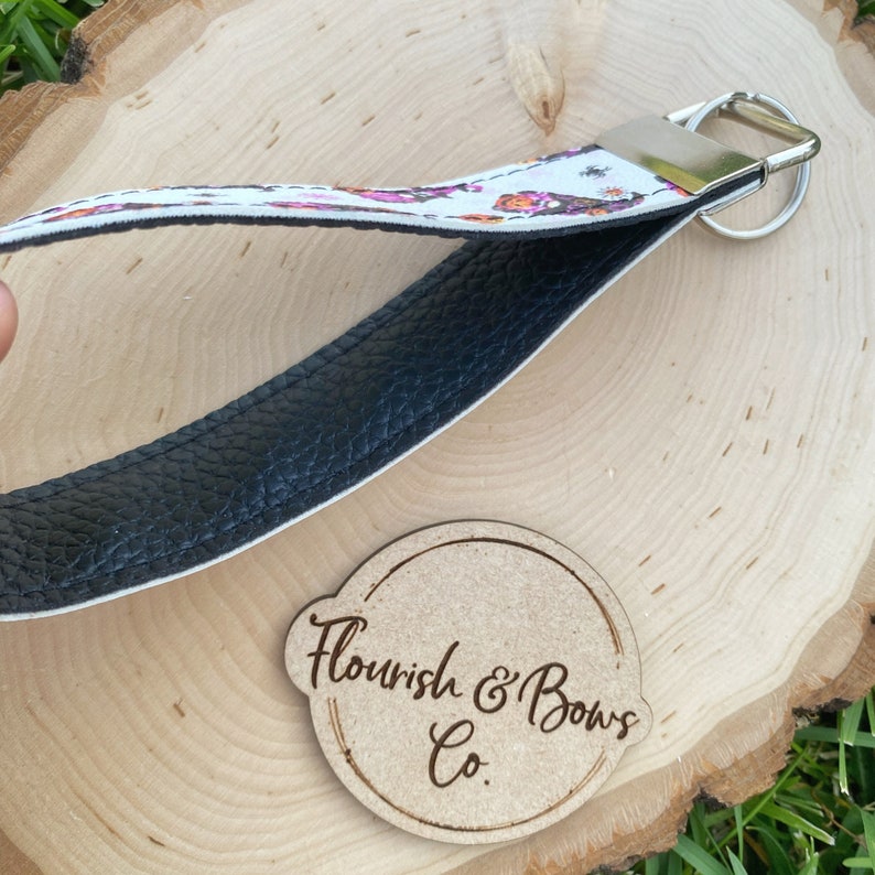 May include: A white key fob with a colorful floral pattern and a black leather interior. The key fob has a silver metal ring and a stitched design. The key fob is from Flourish & Bows Co.