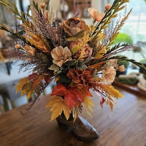 May include: A rustic floral arrangement in a brown cowboy boot vase. The bouquet features a variety of fall-colored flowers, including roses, dahlias, and wheat, with accents of autumn leaves. The boot is made of weathered leather and sits on a wooden table.