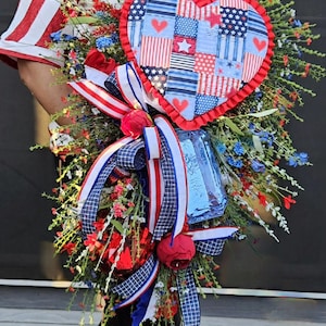 May include: A woman wearing a red and white striped shirt holds a patriotic-themed wreath with a red, white, and blue heart-shaped centerpiece. The wreath is decorated with red, white, and blue ribbons and flowers.