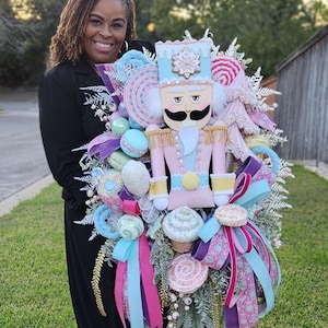 May include: A whimsical Christmas wreath featuring a pastel pink and blue nutcracker figure. The wreath is adorned with colorful candies, cupcakes, ribbons, and decorative elements. The overall design is festive and cheerful.