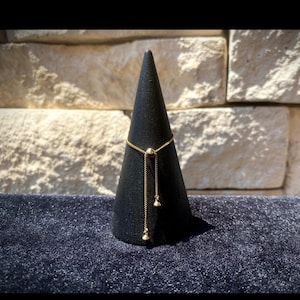 May include: A gold adjustable ring with a small ball and two dangling charms. The ring is displayed on a black cone.