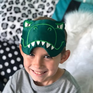 May include: A child wearing a gray t-shirt smiles while wearing a green dinosaur sleep mask.