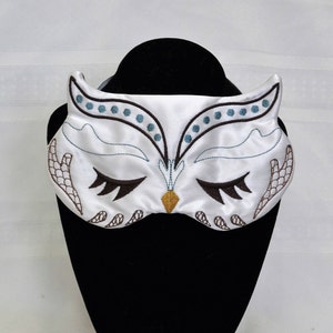 May include: White satin sleep mask with an embroidered owl design. The owl has brown and blue accents and closed eyes.
