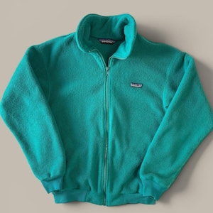 May include: A teal Patagonia fleece jacket featuring a full-length front zipper, a high mock neck collar, and long sleeves with ribbed cuffs. The brand logo patch is stitched onto the left chest, and the fabric has a soft, textured finish.