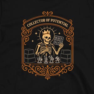 May include: A black short-sleeve t-shirt featuring a graphic print of a skeleton wearing a crown, holding a small grid card, and surrounded by miniature figurines and boxes. The text "COLLECTOR OF POTENTIAL" is arched above the skeleton.