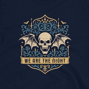 May include: Navy blue t-shirt featuring a skull with bat wings and ornate floral designs in light blue and white. The text "WE ARE THE NIGHT" is displayed on a banner. The shirt is a short-sleeved crew neck.