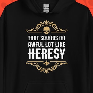 Op de afbeelding: Zwarte hoodie met de tekst "THAT SOUNDS AN AWFUL LOT LIKE HERESY" in wit en goud. Het ontwerp bevat een schedel en decoratieve krullen. De achtergrond is een patroon van rode schedels.