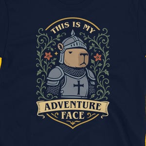 May include: Navy blue t-shirt featuring a cartoon capybara in knight's armor with the text "THIS IS MY ADVENTURE FACE". The design includes floral elements and a banner. The shirt is set against a yellow skull background.