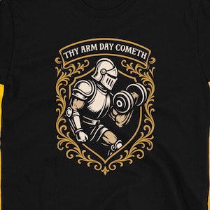 Pode incluir: Camiseta preta com um cavaleiro levantando um haltere dentro de uma moldura dourada ornamentada. O texto "THY ARM DAY COMETH" está acima do cavaleiro. O logotipo Dungeon Arsenal está no canto inferior direito.