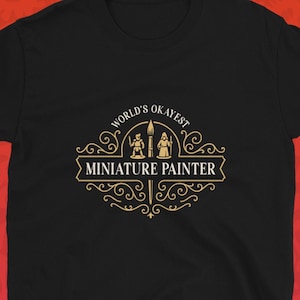 May include: Black t-shirt with a gold and white design that reads "World's Okayest Miniature Painter." The design includes illustrations of a painter and a miniature figure. The Dungeon Arsenal logo is in the bottom right corner.