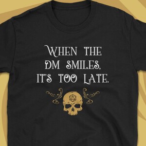 May include: Black t-shirt with the text "When the DM Smiles, It's Too Late." The design includes a gold skull with a D20 die and decorative flourishes. A fun graphic tee for tabletop gamers.