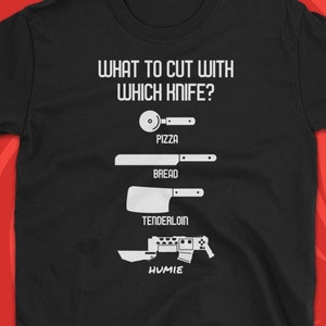 Op de afbeelding: Zwart T-shirt met witte tekst die "What to cut with which knife?" luidt en een reeks messen toont met de labels "Pizza", "Bread", "Tenderloin" en "Humie".