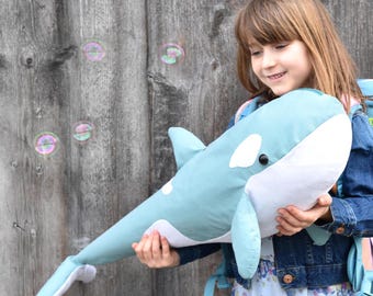 Op de afbeelding: Een jong meisje houdt een blauw en wit pluche orka-walvis speelgoed vast. Ze draagt een blauw denim jack en een bloemenjurk. De orca heeft een witte buik en een zwart oog.