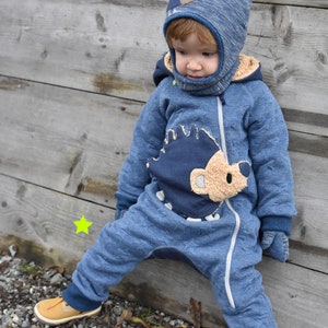 May include: A toddler wearing a blue and brown hedgehog-themed jumpsuit with a matching blue and brown hat. The jumpsuit has a zip down the front and the hat has ear flaps. The toddler is standing on a gravel path with leaves and twigs scattered around.