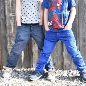 Könnte beinhalten: Zwei Kinder in Jeans stehen vor einem Holzzaun. Ein Kind trägt eine blaue Jeans und ein blaues Shirt mit einem Spiderman-Motiv. Das andere Kind trägt eine dunkelblaue Jeans und ein grau-weißes gepunktetes Shirt.