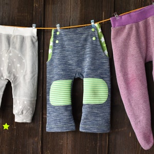 Könnte beinhalten: Drei Paar Babyhosen hängen an einer Wäscheleine. Die Hosen sind grau mit weißen Sternen, blau mit grünen Streifen und lila.