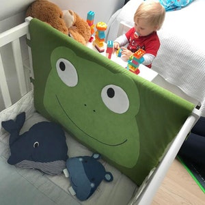 May include: A white crib with a green frog-shaped bumper pad. The crib also has a blue whale and a blue hippopotamus stuffed animal inside. A child is playing with colorful building blocks on a white table next to the crib.