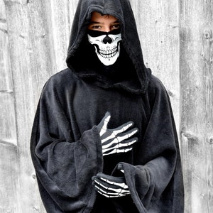 May include: A person wearing a black hooded robe with a skull face mask. The robe has long sleeves and a large hood. The person is standing in front of a wooden wall.