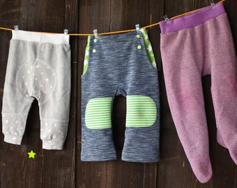 May include: Three pairs of baby pants hanging on a clothesline. The pants are gray with white stars, blue with green stripes, and purple.