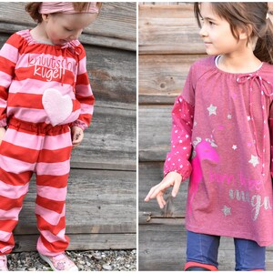 Puede incluir: Dos niñas pequeñas con camisas de manga larga a rayas rojas y rosas. La niña de la izquierda lleva un mono a rayas rojas y rosas con un corazón rosa en la parte delantera. La niña de la derecha lleva una camisa de manga larga rosa con un unicornio y estrellas impresas en la parte delantera. Ambas niñas llevan pantalones vaqueros.