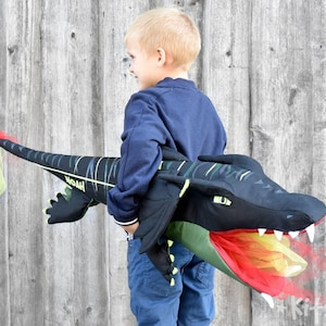 May include: A child wearing a blue shirt holds a black and green dragon puppet with a red and yellow fire mouth. The dragon has a green and yellow stripe pattern and the text "Woah" is visible on the side.